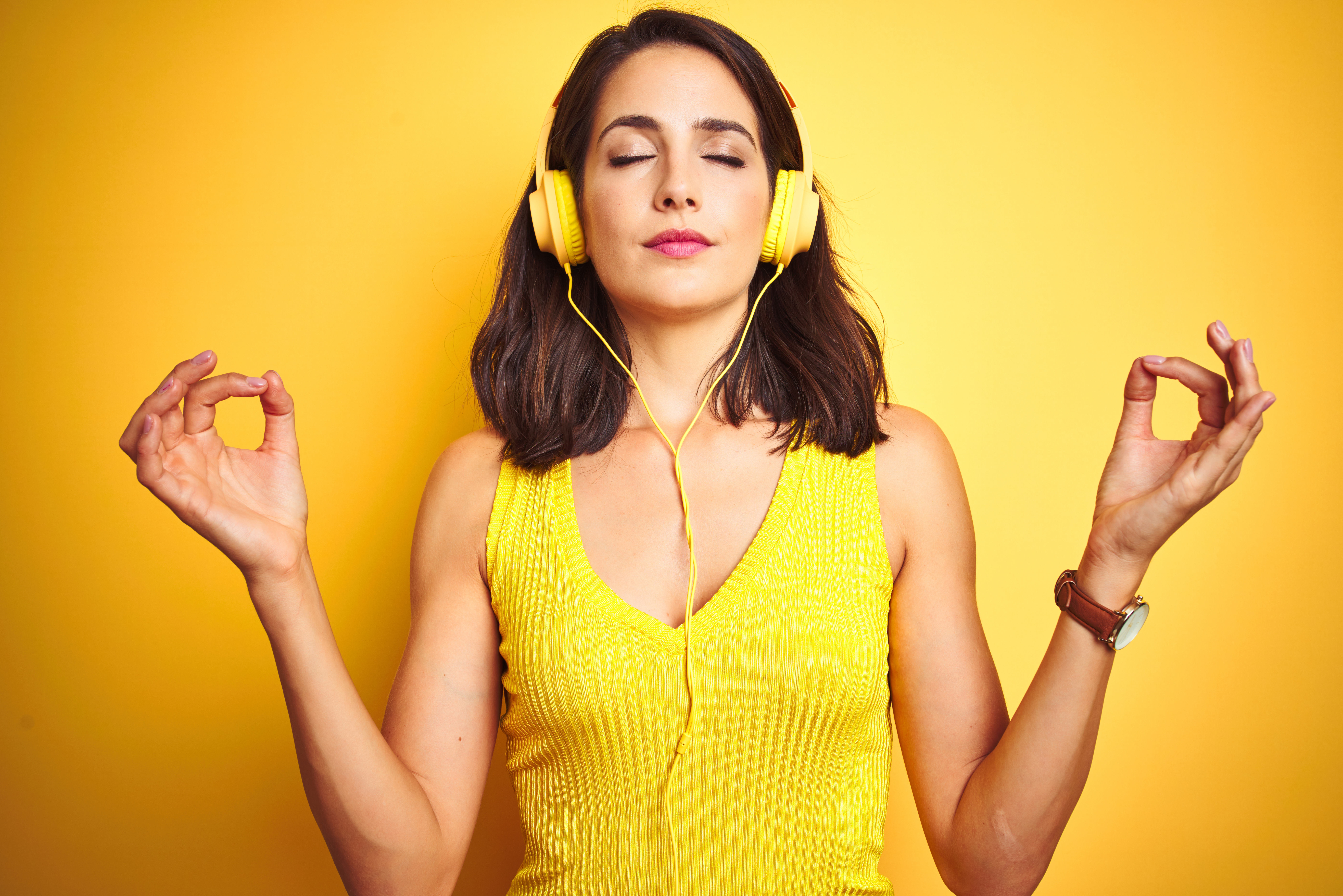 Young beautiful woman listening to music using headphones over yellow isolated background relax and smiling with eyes closed doing meditation gesture with fingers. Yoga concept.